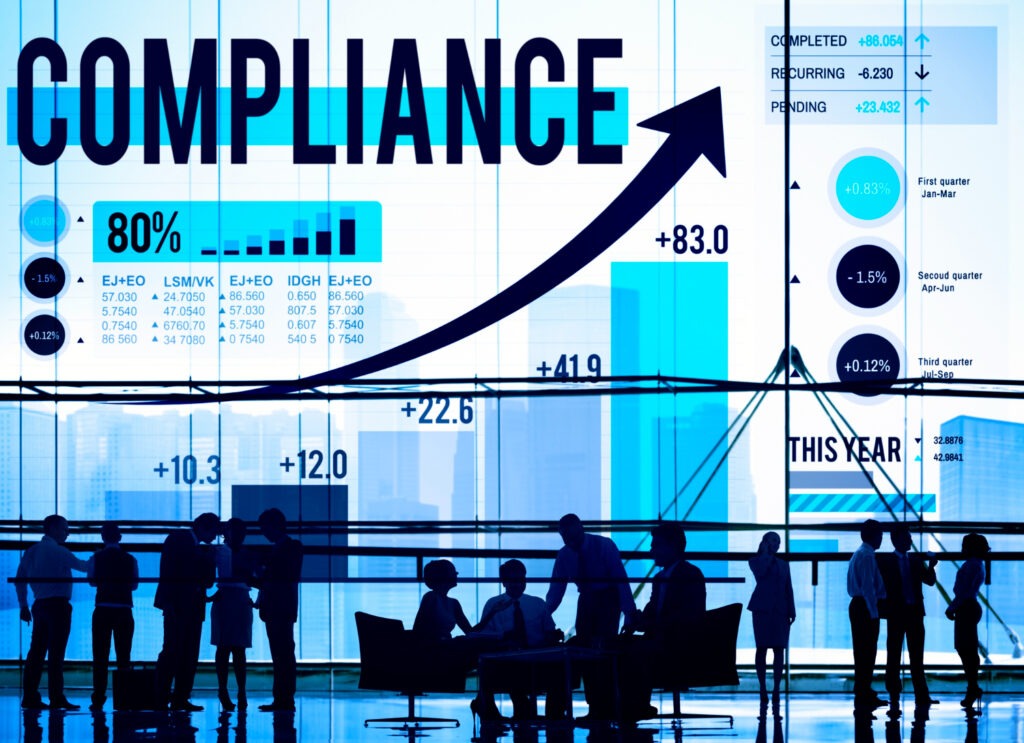 A business-themed image with the word 'COMPLIANCE' in bold, overlaid with charts, graphs, and financial statistics. Silhouettes of professionals in discussion appear in the background